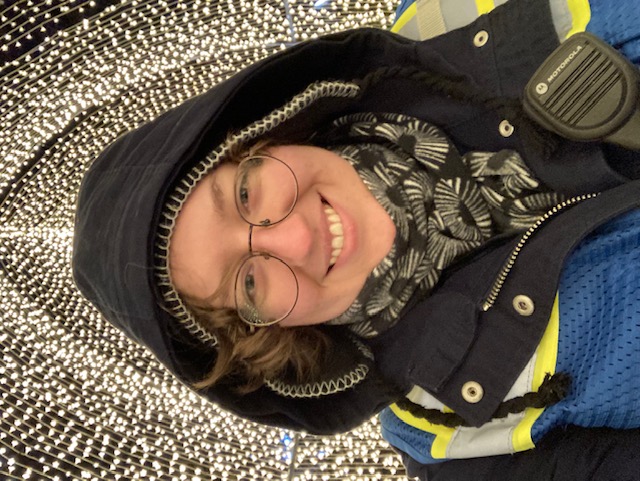 A headshot of Maggie working at the Brookyln Botanic Gardens's winter show, Lightscape. She is bundled up for the winter in a hat, scarf, and coat with the hood pulled over her head. She also wears a reflective vest and a walkie talkie, and is smiling under warm, cascading lights.